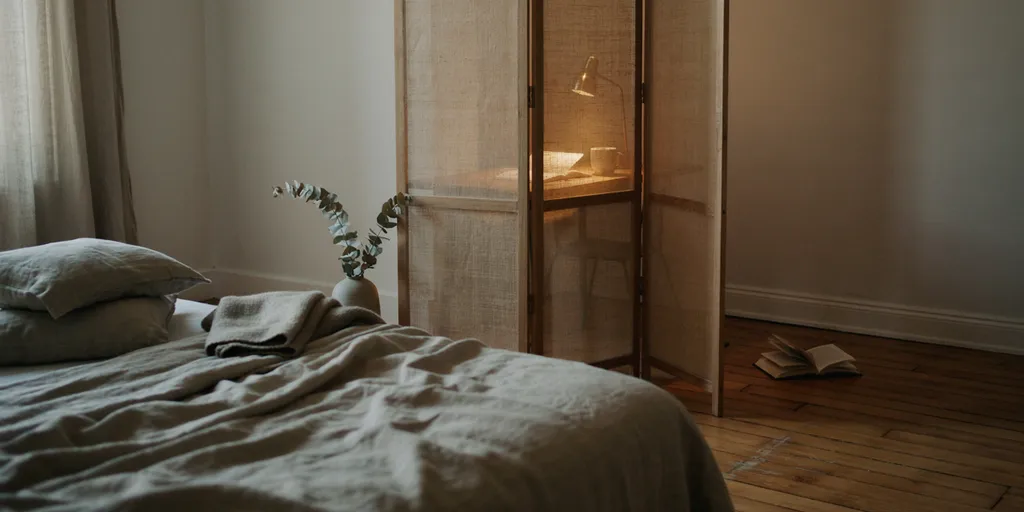 A bedroom where a folding linen screen stands closed in front of a small desk, keeping the work out of sight from the bed