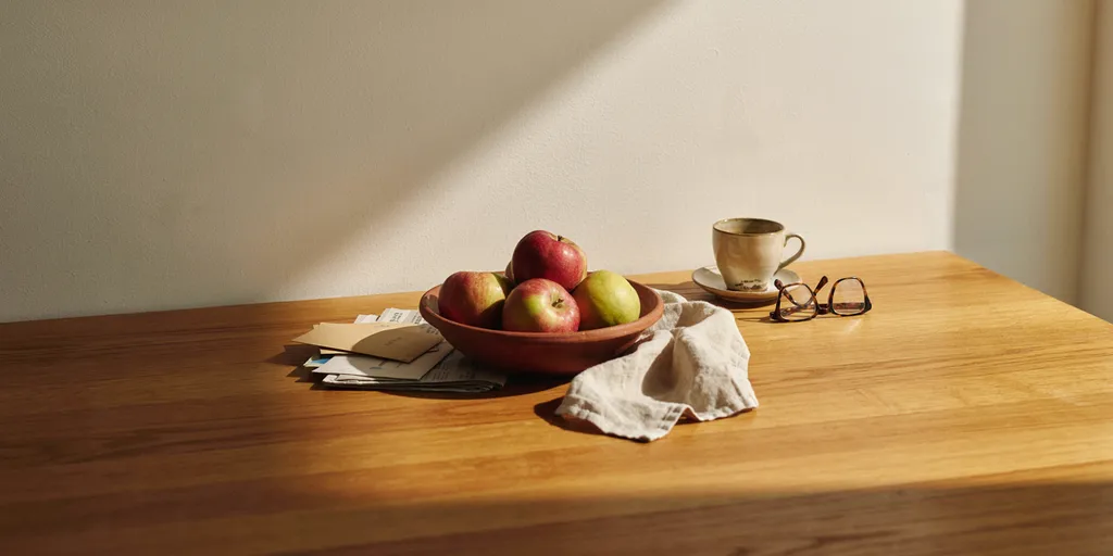 An oiled oak table with a clay bowl of apples, a small pile of post, a half-drunk cup and reading glasses, every other surface clear