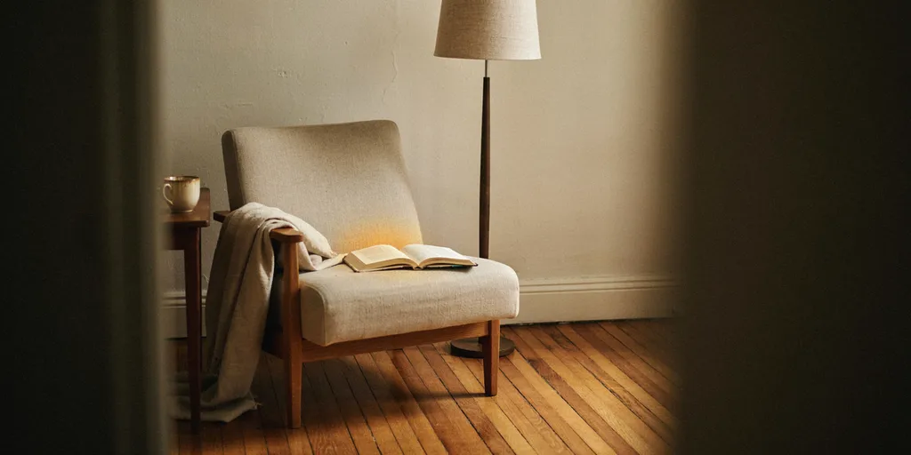 A tall floor lamp behind and slightly to the side of a reading chair, its light falling forward onto an open book without casting a shadow on the page