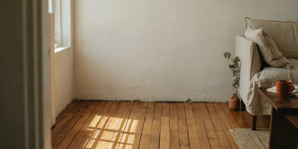 Two plaster walls meeting above bare swept oak floorboards with the corner left completely empty, a linen sofa at the edge of the frame