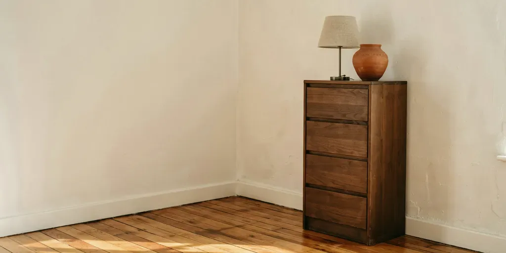 A tall narrow walnut chest of drawers in the corner of a small bedroom, a lamp and a clay vase on top