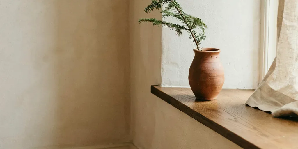 A single sprig of evergreen in a small clay bud vase on a bedroom windowsill in soft morning light